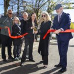 Six people stand in a roadway in the center of the frame with two parts of a red ribbon held by people on either side. A person in the center holds a pair of oversized golden scissors. A green, metallic arch stands over the right side of the frame, and trees with green leaves stand on the left side of the roadway.