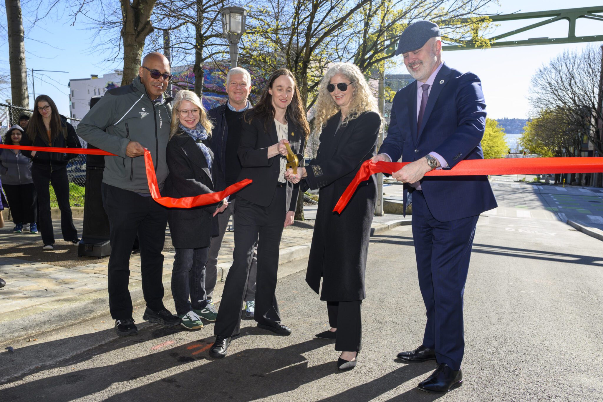 Six people stand in a roadway in the center of the frame with two parts of a red ribbon held by people on either side. A person in the center holds a pair of oversized golden scissors. A green, metallic arch stands over the right side of the frame, and trees with green leaves stand on the left side of the roadway.