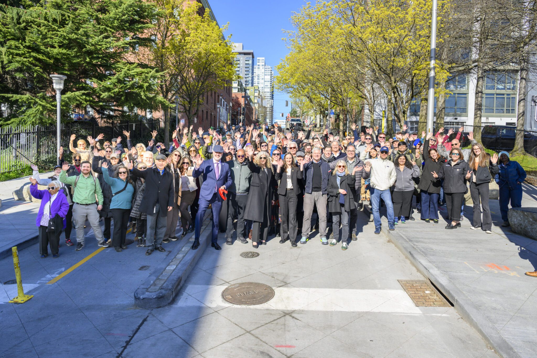 A large group of people stand waving together in the center of a roadway and on the adjacent sidewalks on either side. Trees with green leaves line the sides of the street, and the buildings on either side of the roadway become progressively taller in the distance. 