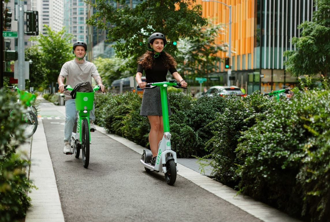 A woman rides a scooter and a man rides a bike in a separated bike lane.