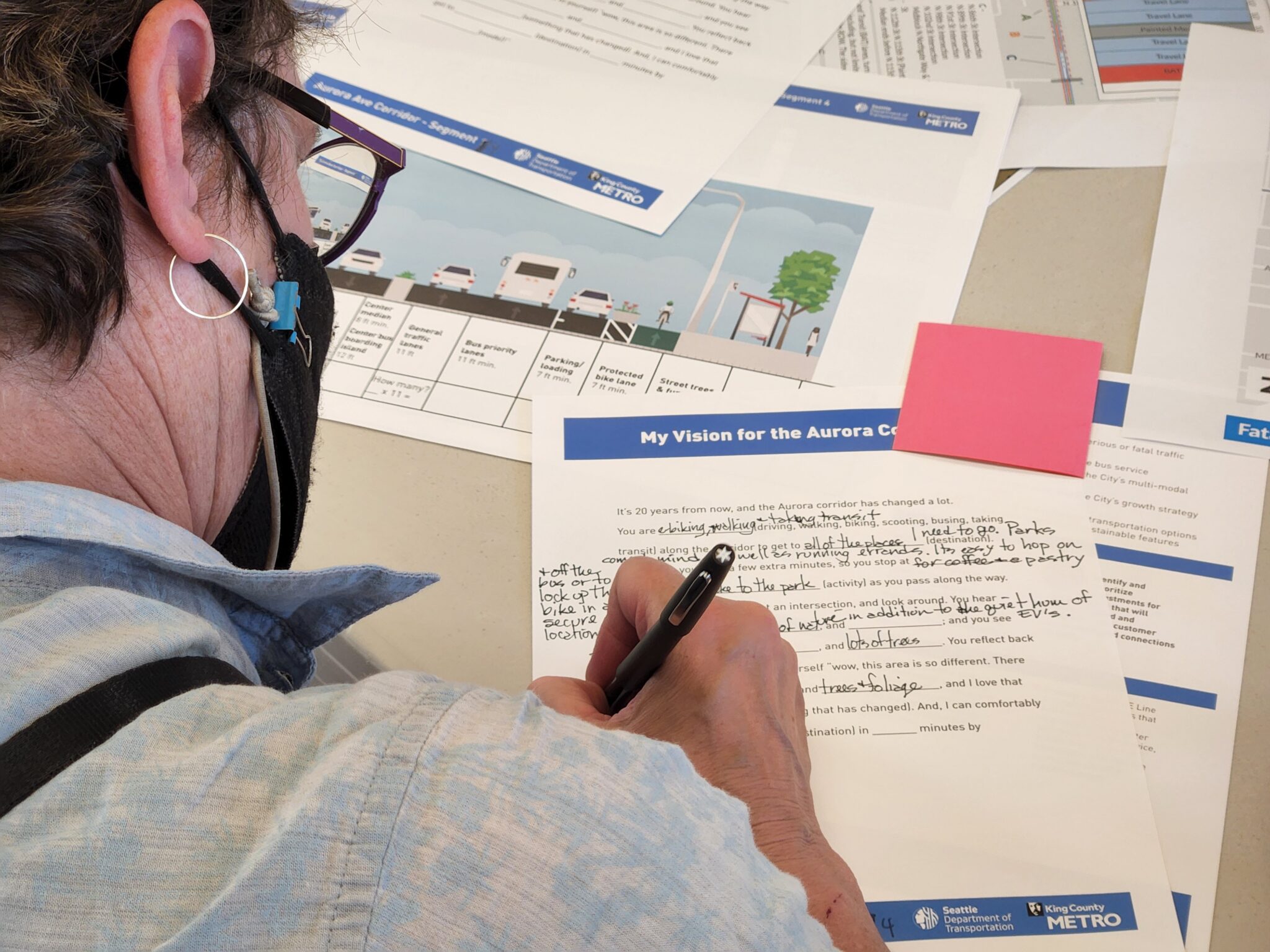A person with glasses and a black face mask is writing on a feedback form. The table is covered with planning documents. Logos for SDOT and King County Metro are visible on the bottom of the form. The top of the form says 