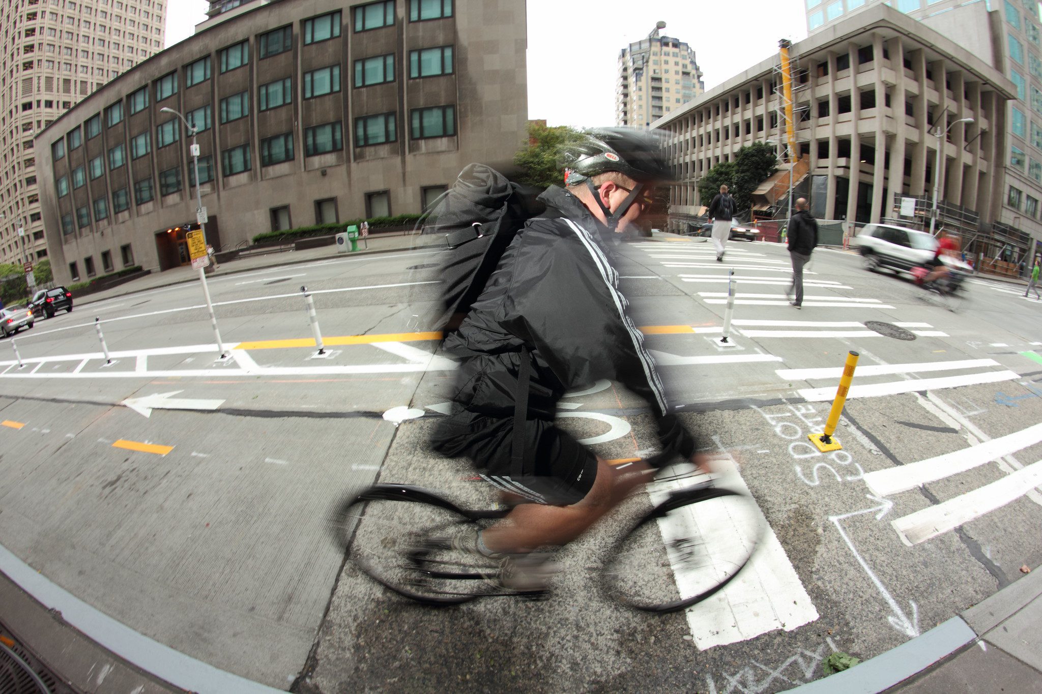A cyclist in motion blurs across an urban intersection inside a protected bike lane surrounded by tall buildings. The cityscape with marked lanes, bollards, and crosswalks. Fisheye distortion adds depth and focus to the cyclist as the central subject.