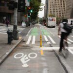 An urban street features a protected bicycle lane with bright green markings, a blurred cyclist in motion, and a FedEx truck driving away. Traffic signals, pedestrians, and a mix of safety elements like bollards and crosswalks highlight the integration of bike-friendly infrastructure in a busy cityscape.