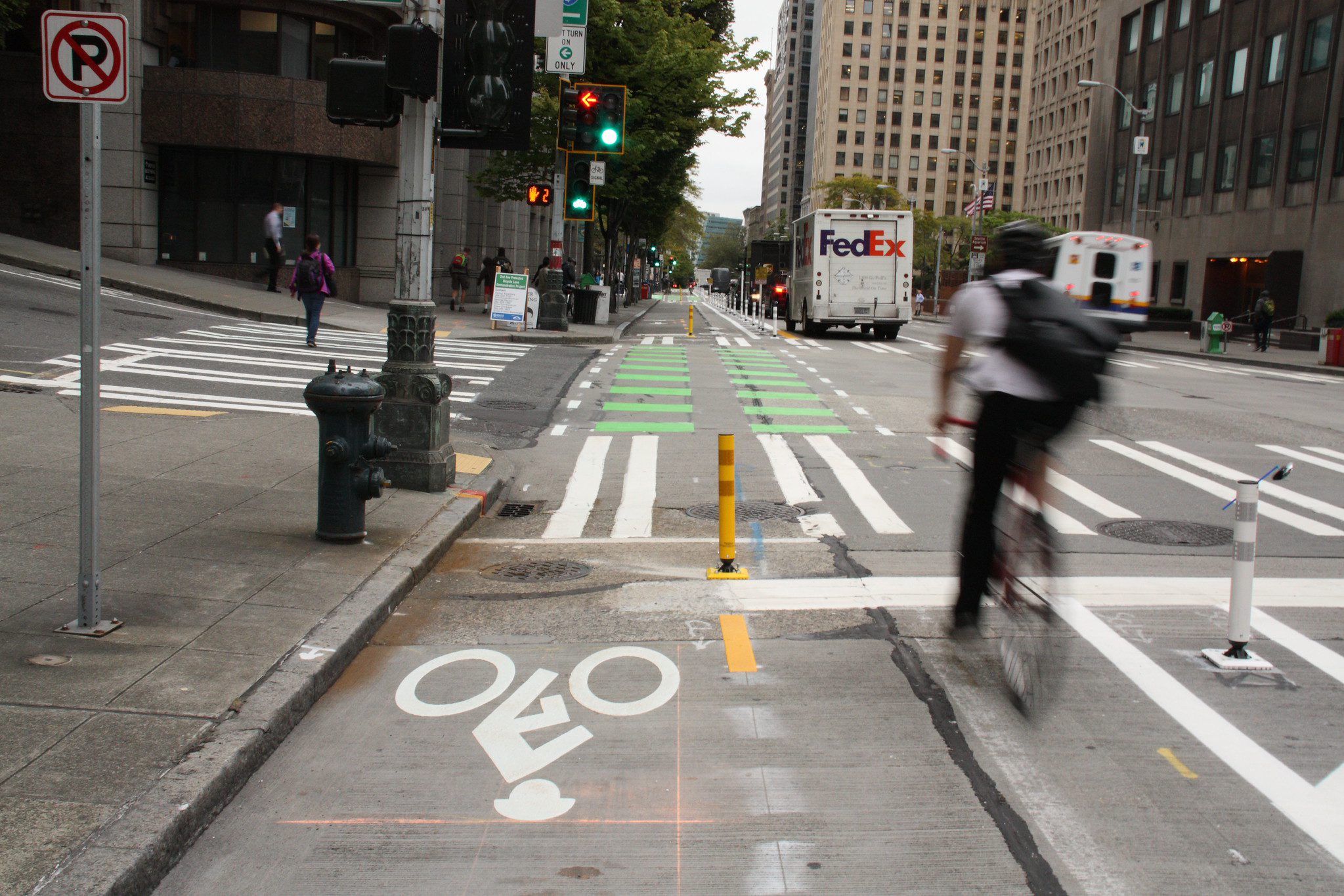 An urban street features a protected bicycle lane with bright green markings, a blurred cyclist in motion, and a FedEx truck driving away. Traffic signals, pedestrians, and a mix of safety elements like bollards and crosswalks highlight the integration of bike-friendly infrastructure in a busy cityscape.