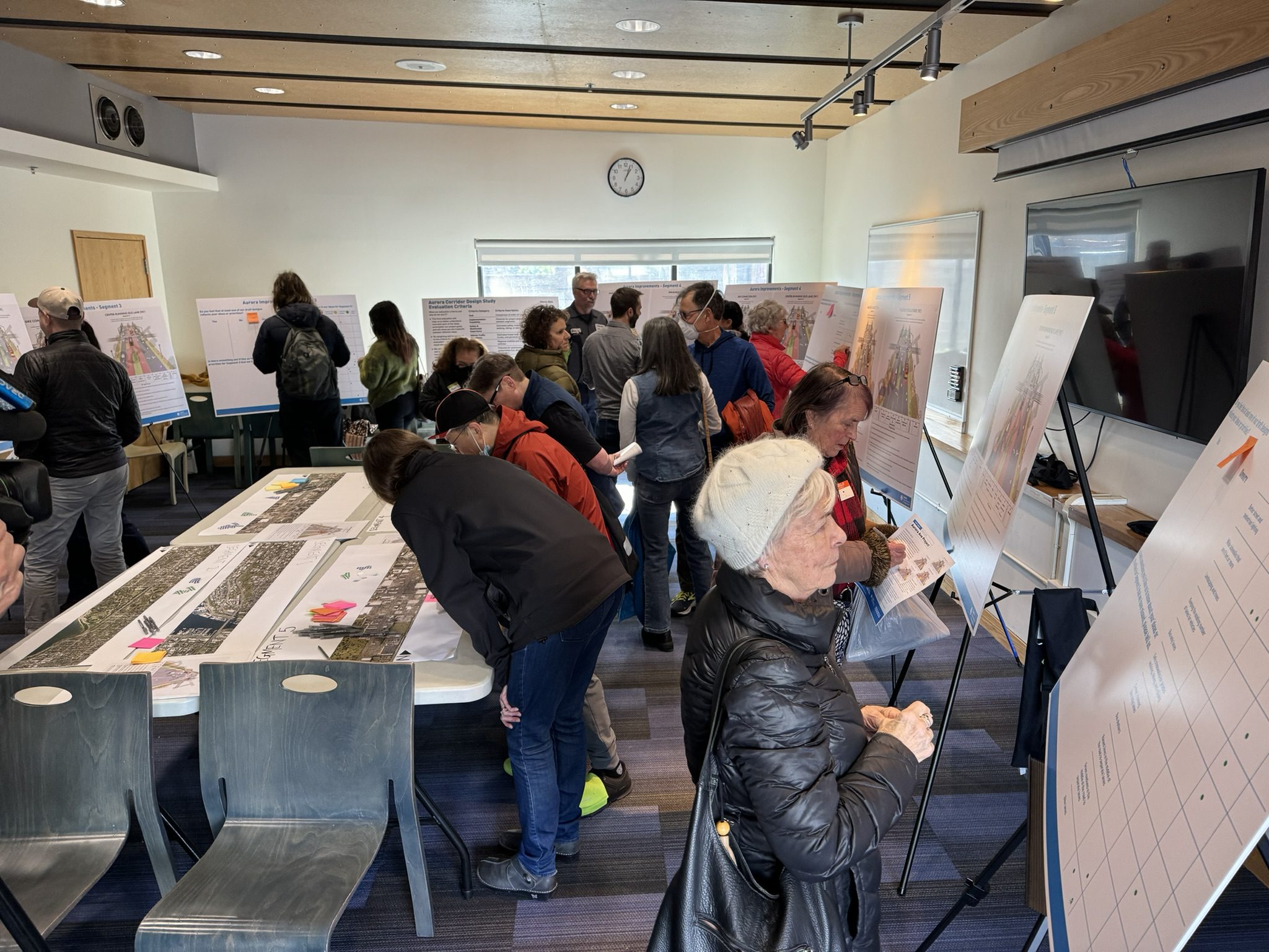An indoor community meeting. 15-20 people review maps and posters. The room features white walls, a wood-paneled ceiling, and blue-gray carpet. 