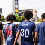Four people are walking toward a large sports stadium, viewed from behind. Each wears a distinct jersey. From left to right: a black jersey with a gold the number "10," a shiny blue jersey with "OKOCHA" and "10" in red and white, a navy blue jersey with "MESSI" and "30" in white, and a matte blue jersey with a subtle dark "10." The background features a modern stadium with a white arched roof, a tall grey tower with circular icons (a clock, a yellow gear, and a green circle), lush green trees, and a paved walkway