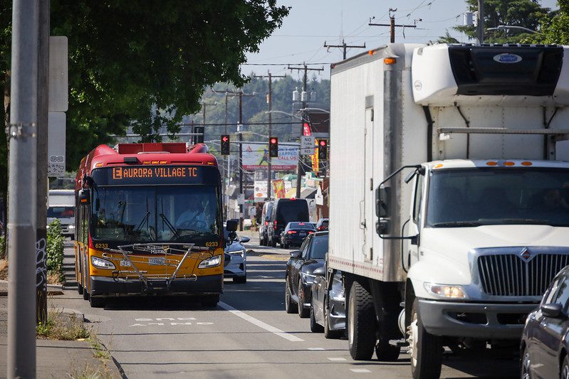 A red and yellow bus is driving in a 'Bus Only' lane on a busy urban street. The bus displays 'E Aurora Village TC' on its destination sign. To the right, a large white freight truck and several cars are queued in traffic. The sky is clear and bright, suggesting a sunny day.
