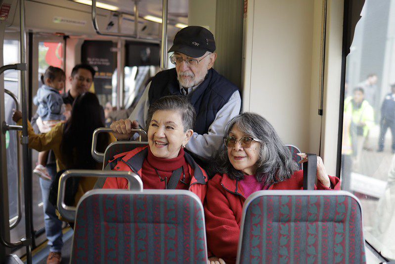 Two women sit next to each other, smiling, inside a streetcar. A man stands behind them. Further in the distance, another man boards the streetcar holding a child. 