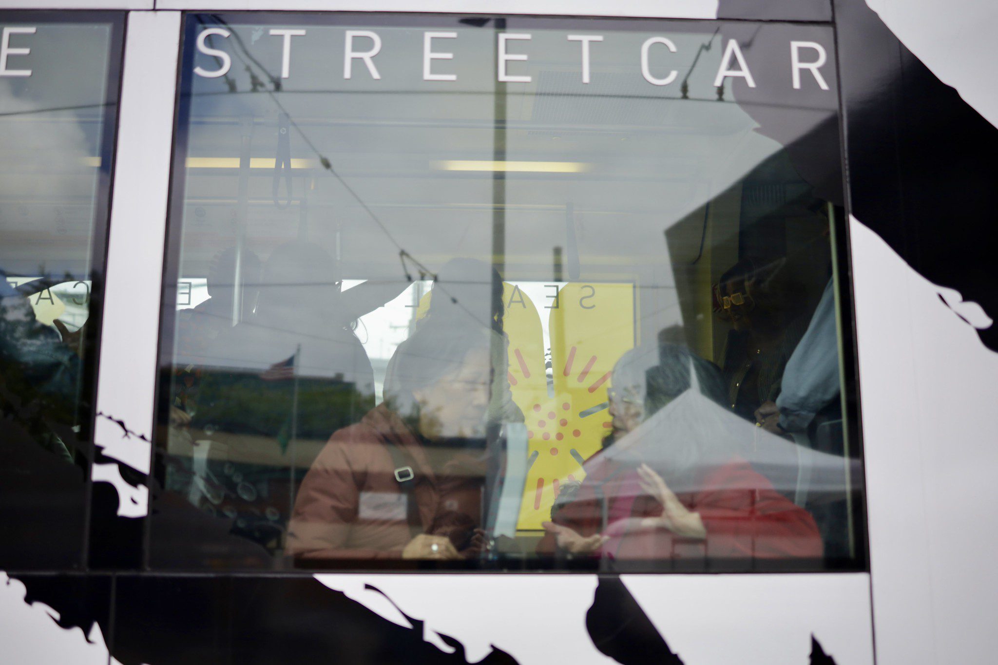 An outside view of a window with the word "STREETCAR" on the top of it. Through the glass two women sit talking to each other. Outlines of people standing can been seen in the distance.