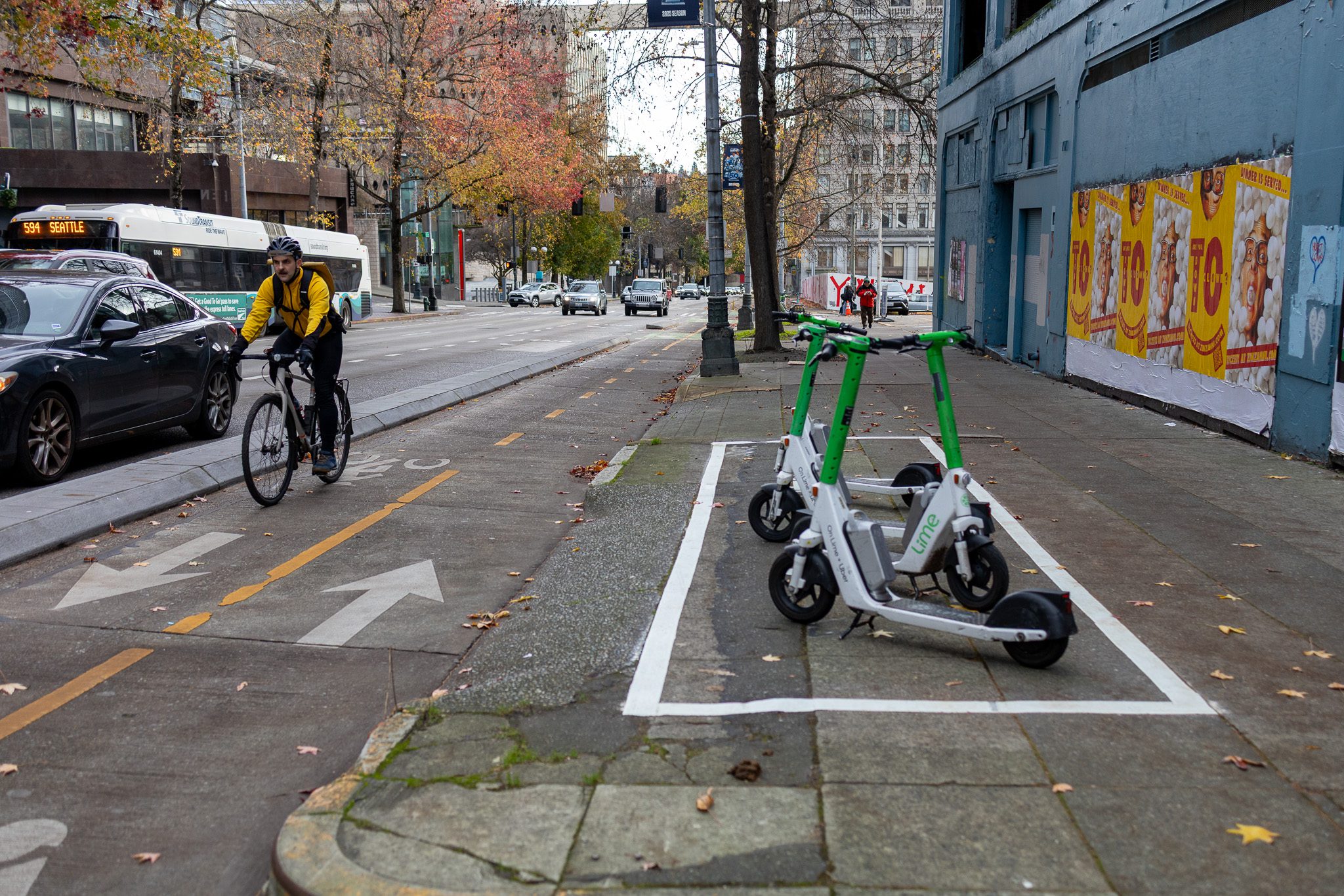 A person wearing a yellow jacket and black helmet rides a bike inside a bike lane. To the right, four green and white electric scooters are parked on the sidewalk. The ground has white markings making a rectangle, where the scooters are parked inside.
