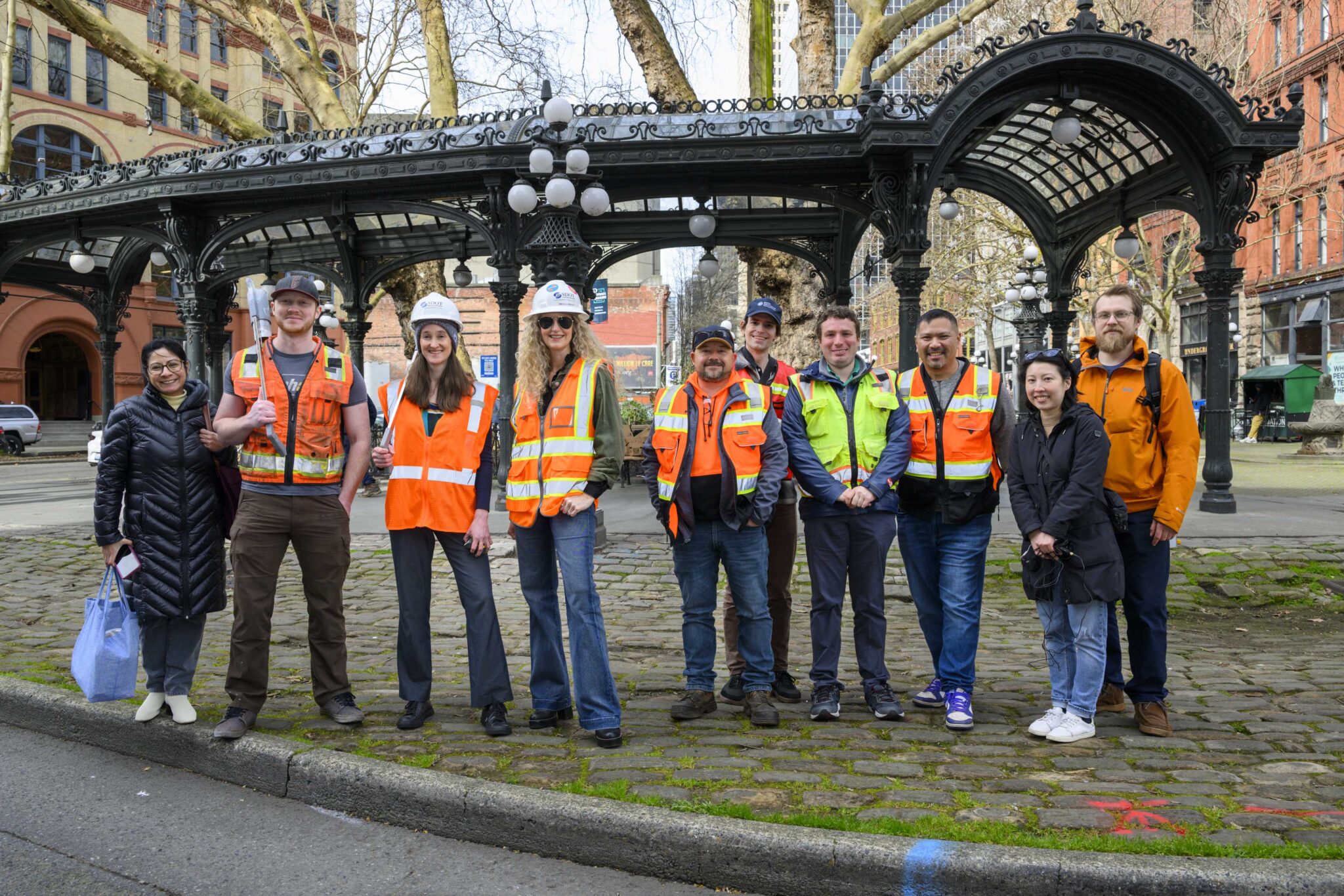 A group of ten people, wearing safety vests and hard hats, stand smiling in an urban outdoor setting. The image looks identical to the previous photo but the cartoon water imagery and fish puppet are gone.  The people are standing on cobblestone. 
