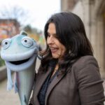 A puppet in the from of a fish and a women smile at each other. Trees and sidewalks are in the background.