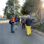 A group of about eight people gathers on a residential street corner under bright sunlight, dressed in cold-weather clothing. The scene includes a man in a high-visibility orange vest, suggesting an organized community event, with participants standing in a semi-circle near trees, parked cars, and a sloping embankment.