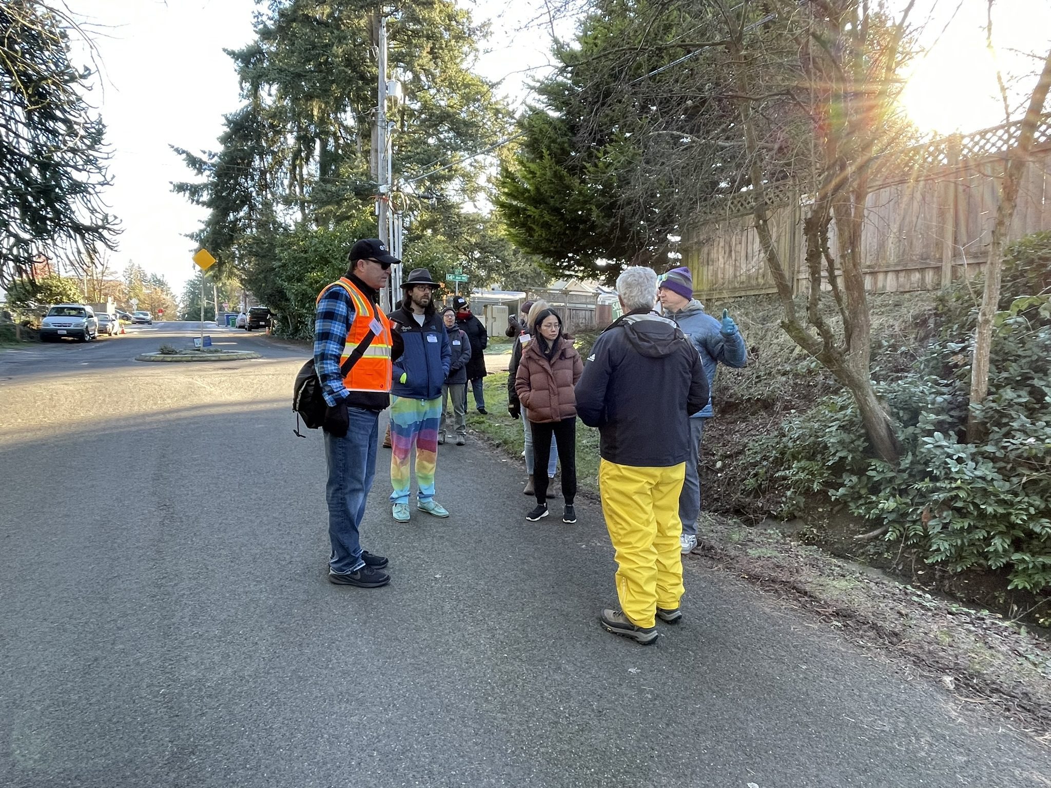A group of about eight people gathers on a residential street corner under bright sunlight, dressed in cold-weather clothing. The scene includes a man in a high-visibility orange vest, suggesting an organized community event, with participants standing in a semi-circle near trees, parked cars, and a sloping embankment.