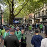 ALT TEXT: A large crowd of people, many wearing bright green and blue jerseys, fill a brick-paved street in Pioneer Square. They are surrounded by historic buildings and leafy green trees. A stage displays the slogan "ONE TEAM. ONE GOAL."