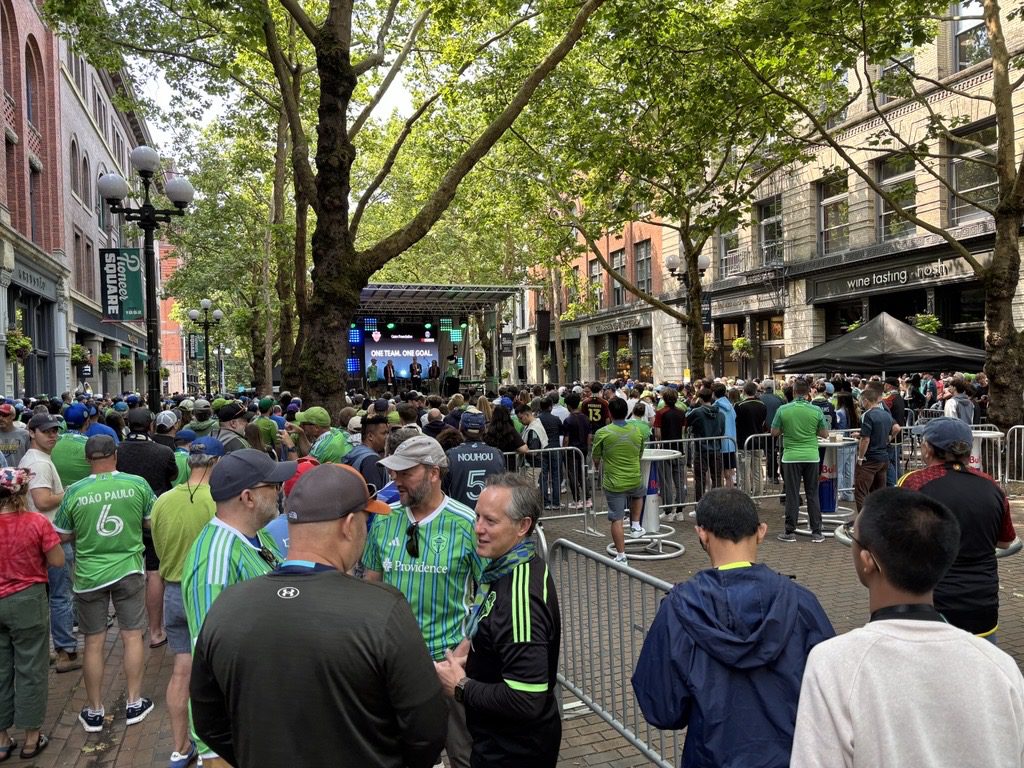ALT TEXT: A large crowd of people, many wearing bright green and blue jerseys, fill a brick-paved street in Pioneer Square. They are surrounded by historic buildings and leafy green trees. A stage displays the slogan 