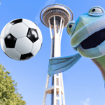 A fish puppet smiles in front of the iconic Space Needle with a soccer ball floating mid-air nearby.