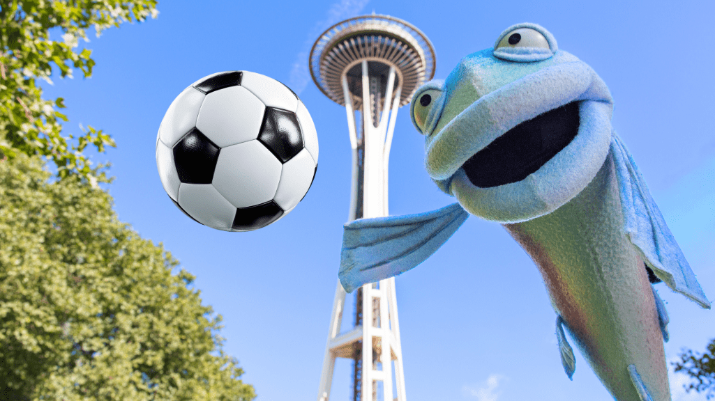 A fish puppet smiles in front of the iconic Space Needle with a soccer ball floating mid-air nearby.