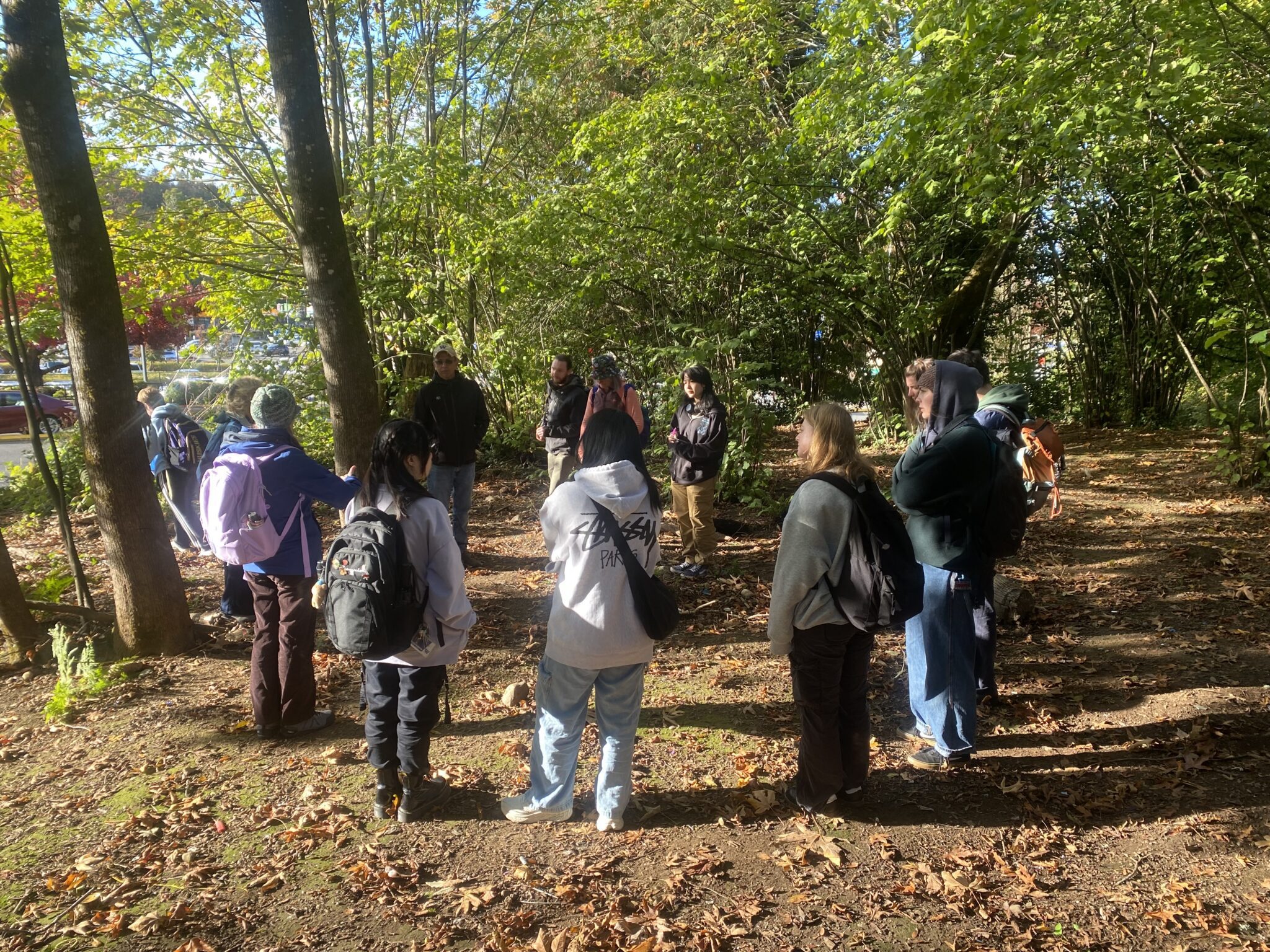 A group of about 12 young people wearing backpacks gather in a wooded area on a sunny day. They stand in a loose circle, focused on a man in a black jacket and baseball cap who appears to be leading the group. Sunlight filters through the dense green tree canopy. 
