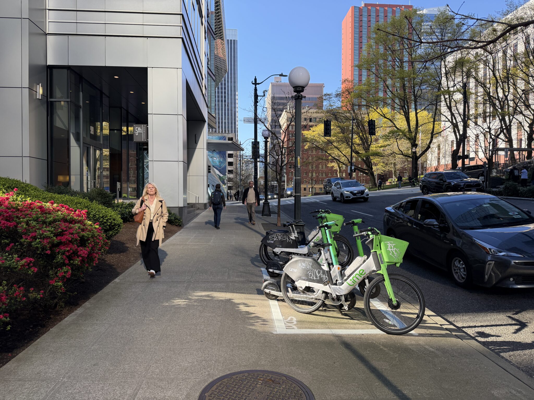 A sunny urban scene featuring a woman in a tan trench coat walking on a wide sidewalk. To the right, whit markings on the sidewalk create a rectangle on the ground. White and green electric bicycles are parked inside the rectangle. 