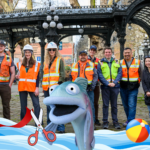 Alt text: A group of ten people, wearing safety vests and hard hats, stand smiling in an urban outdoor setting. Playful water-themed graphics, including a fish puppet, scissors, and a ribbon, overlay the bottom of the image, suggesting a celebratory event. The scene hints at a ribbon-cutting for a cartoon body of water.