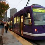 A purple streetcar is stopped at a city platform at dusk, with its interior lights glowing and passengers boarding near the open door. The sign on the front says "Seattle Streetcar" in bright yellow lights. Motion-blurred pedestrians walk along the platform beside autumn-colored trees, while a city skyline rises in the background under a gray evening sky.
