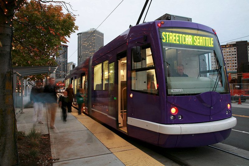 A purple streetcar is stopped at a city platform at dusk, with its interior lights glowing and passengers boarding near the open door. The sign on the front says 