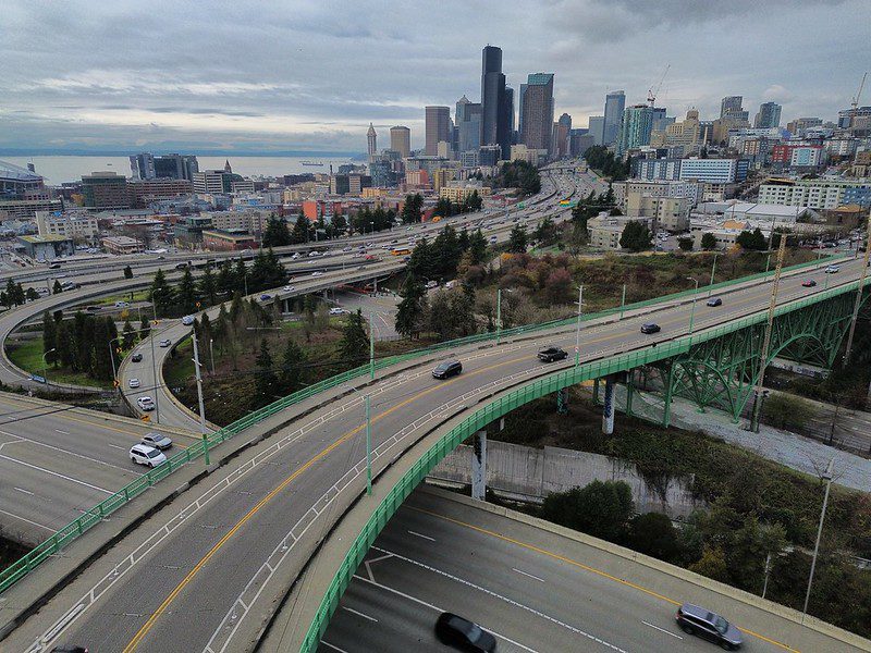 A sky view of a city shows a large green arched bridge crossing above a busy multi-lane highway interchange, with cars moving along curving ramps below. In the distance, Seattle’s downtown skyline rises under a gray, overcast sky, with patches of trees and hints of waterfront adding depth to the urban scene.