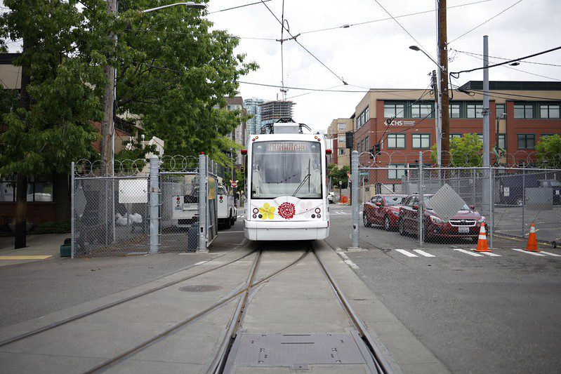 A white First Hill Streetcar faces the camera on tracks leading through a fenced urban area under an overcast sky. The streetcar’s front display reads “Jackson Street,” and colorful floral decals brighten its front, while parked cars, traffic cones, trees, and city buildings fill the background.