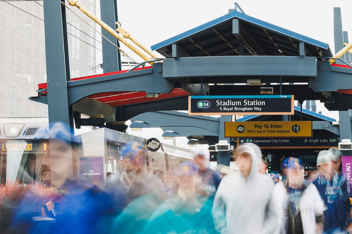 A busy crowd moves through Seattle’s Stadium Station while a light rail train waits at the platform under large steel beams and station signs. The people in the foreground are blurred with motion, while the train and bright directional signs remain sharp in the background.
