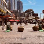 A busy pedestrian street at Seattle’s Pike Place Market is blocked to car traffic with planters, barricades, and road signs while shoppers and visitors walk between storefronts and outdoor seating. A white delivery van is parked along the right side, and tall modern skyscrapers rise behind the historic market buildings under a clear blue sky.
