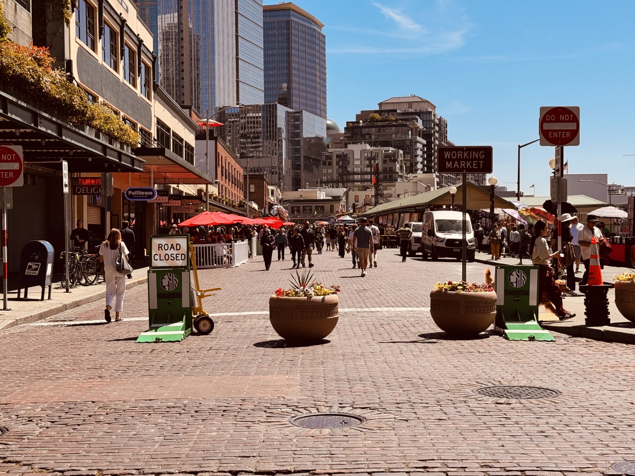 A busy pedestrian street at Seattle’s Pike Place Market is blocked to car traffic with planters, barricades, and road signs while shoppers and visitors walk between storefronts and outdoor seating. A white delivery van is parked along the right side, and tall modern skyscrapers rise behind the historic market buildings under a clear blue sky.