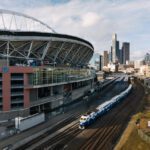 A train travels along the tracks beside a sports stadium. The stadium’s sweeping roof fills the left side of the scene with downtown skyscrapers rising in the background. The photo is taken from an elevated viewpoint on a bright day, showing roads, cars, and trees that frame the busy urban corridor between the stadium and the city.