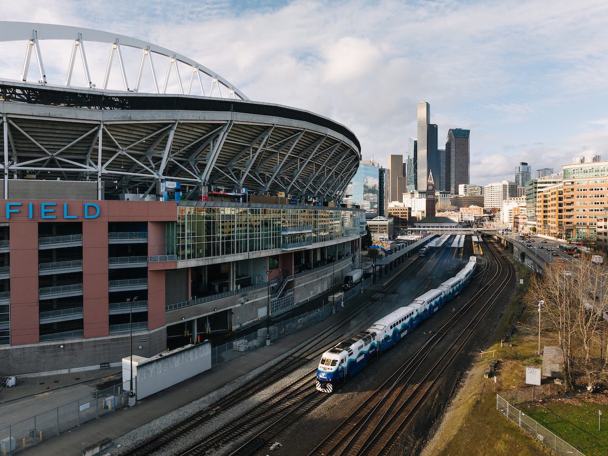 A train travels along the tracks beside a sports stadium. The stadium’s sweeping roof fills the left side of the scene with downtown skyscrapers rising in the background. The photo is taken from an elevated viewpoint on a bright day, showing roads, cars, and trees that frame the busy urban corridor between the stadium and the city.