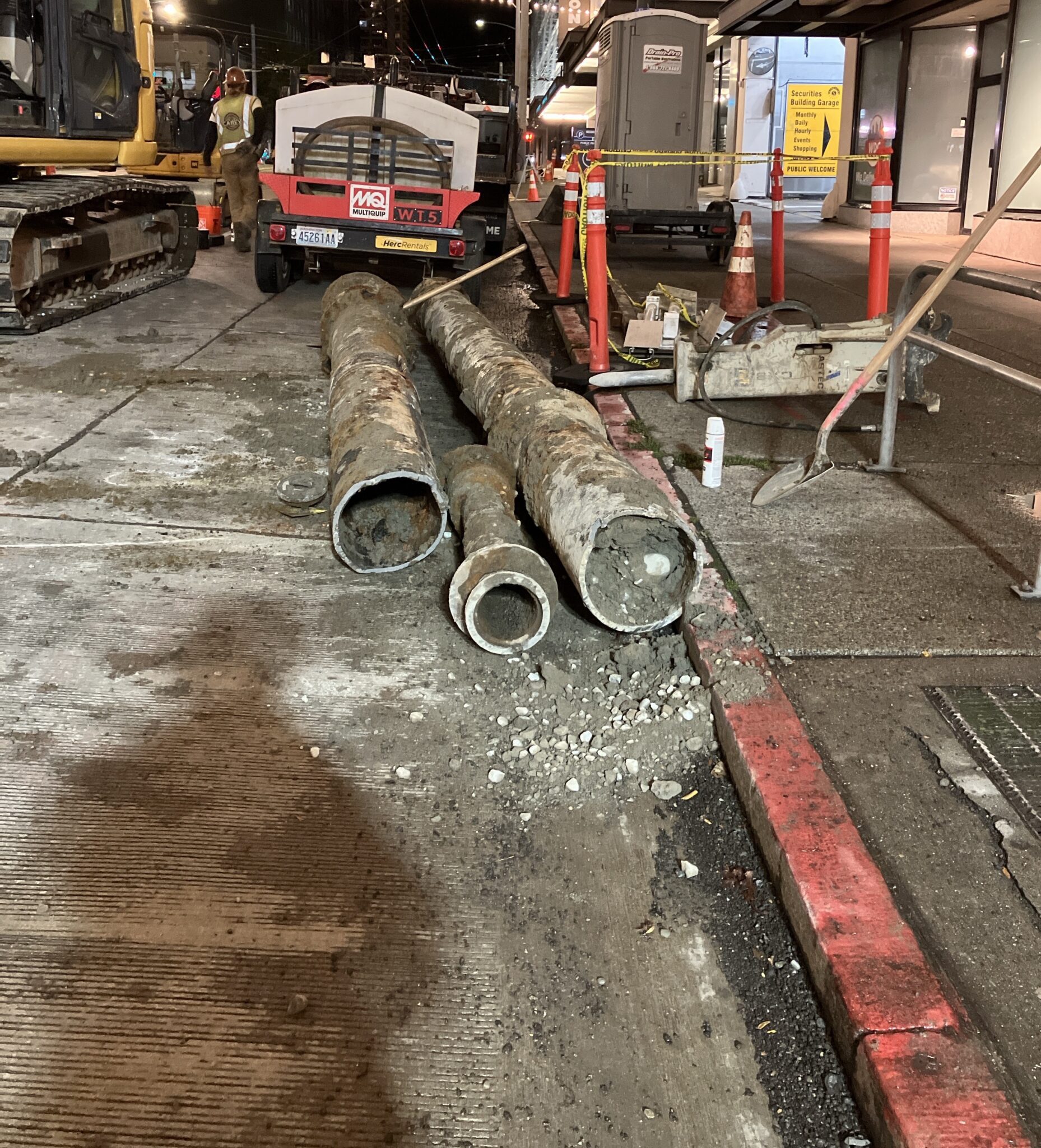 Large rusted pipe sections lie on the ground at the center of a nighttime construction site, surrounded by gravel, heavy equipment, and safety cones.