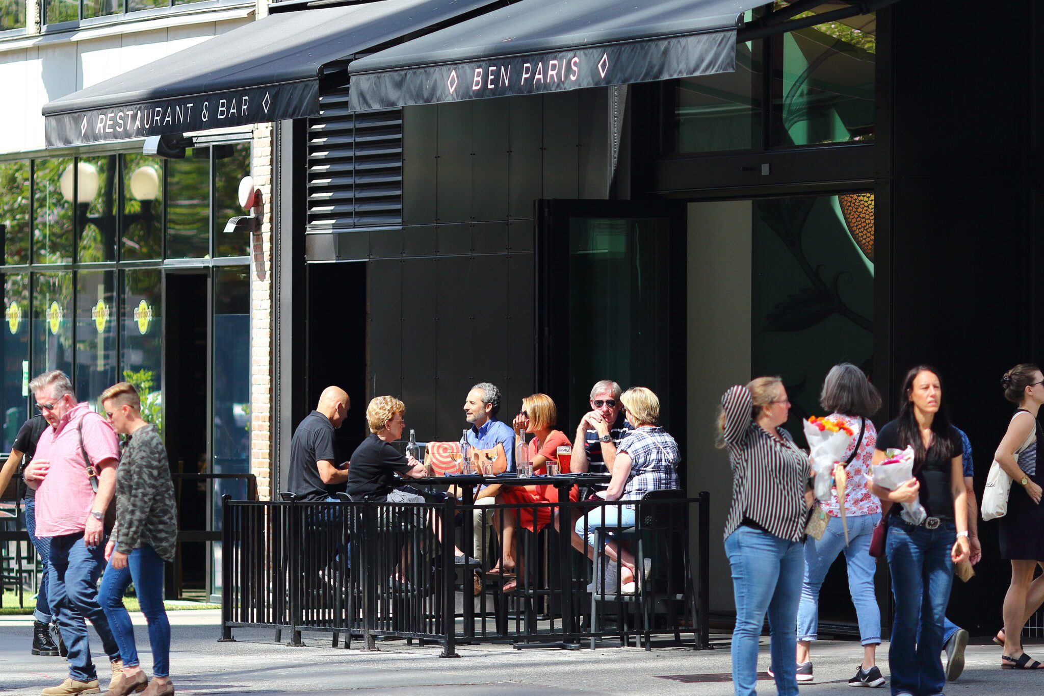 A group of people sits together at an outdoor restaurant patio while several pedestrians pass by on the sidewalk in front of them. The dark awnings and black exterior frame the scene, with bright sunlight casting strong shadows across the street.