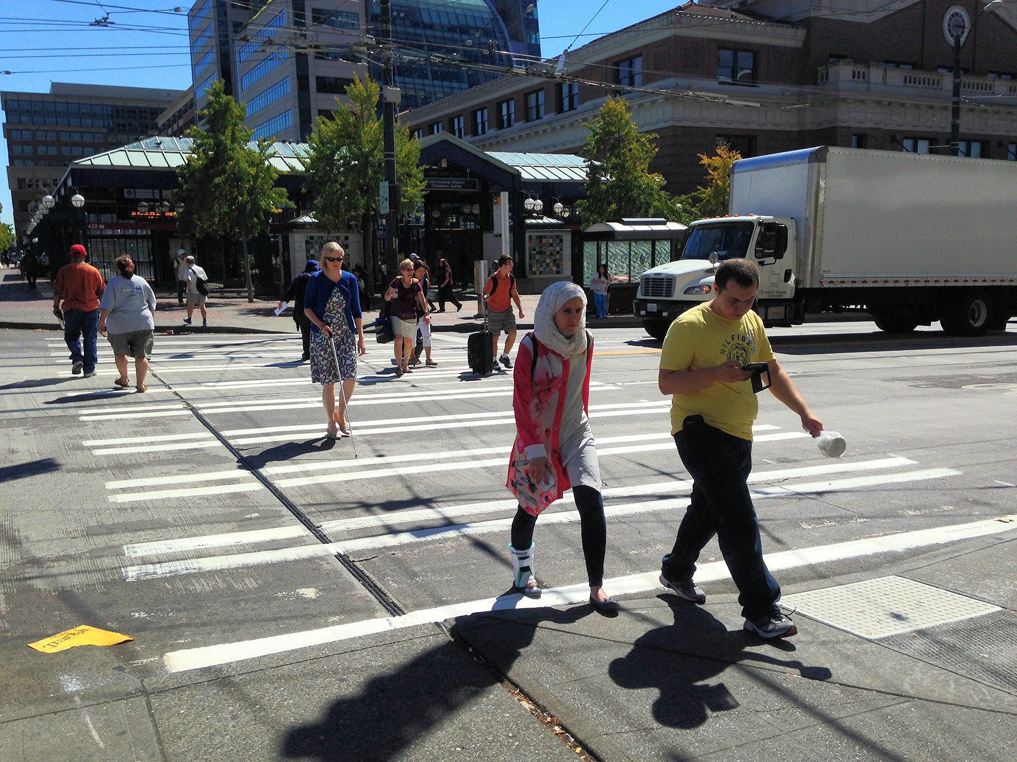 A busy city crosswalk filled with pedestrians moving in different directions on a bright, sunny day. In the foreground, a man in a yellow shirt looks at his phone while walking beside a woman in a white headscarf, and farther back another woman crosses with a white cane as a large white truck passes through the intersection behind them.