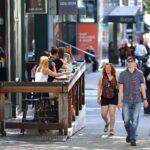 A busy city sidewalk scene shows people dining at an outdoor sushi restaurant while pedestrians walk past in the foreground. A man in a striped polo and a woman with red hair are closest to the camera, with more people and storefronts stretching into the background.