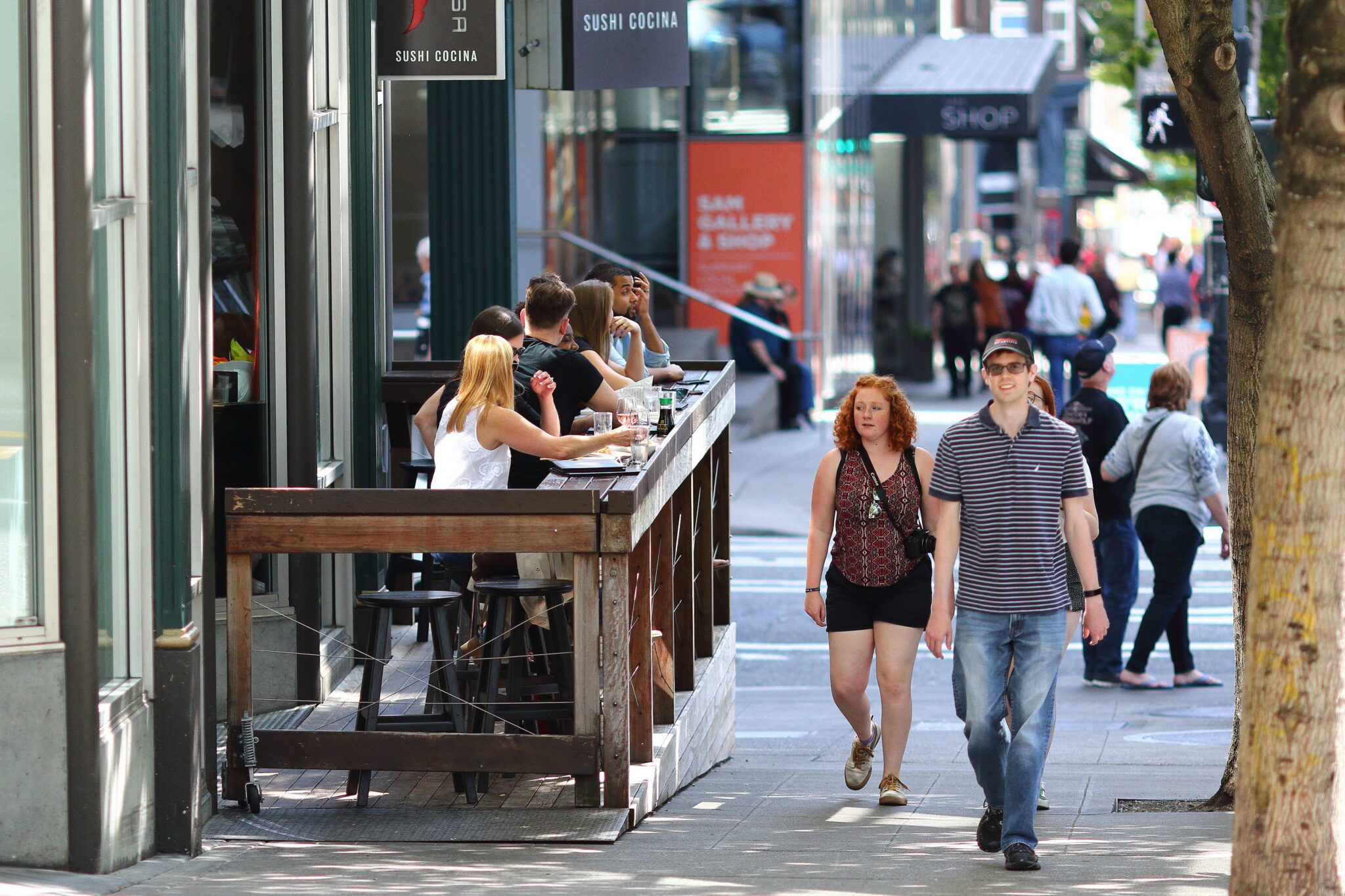 A busy city sidewalk scene shows people dining at an outdoor sushi restaurant while pedestrians walk past in the foreground. A man in a striped polo and a woman with red hair are closest to the camera, with more people and storefronts stretching into the background.