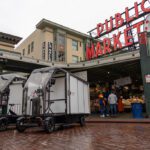 Two electric cargo delivery vehicles are parked on a wet brick street in front of Seattle’s iconic Pike Place Market sign on a gray, rainy day. A few people stand near the market entrance, while the bright red neon sign and clock rise above the scene, contrasting with the muted buildings and overcast sky.