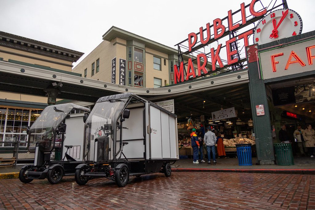 Two electric cargo delivery vehicles are parked on a wet brick street in front of Seattle’s iconic Pike Place Market sign on a gray, rainy day. A few people stand near the market entrance, while the bright red neon sign and clock rise above the scene, contrasting with the muted buildings and overcast sky.