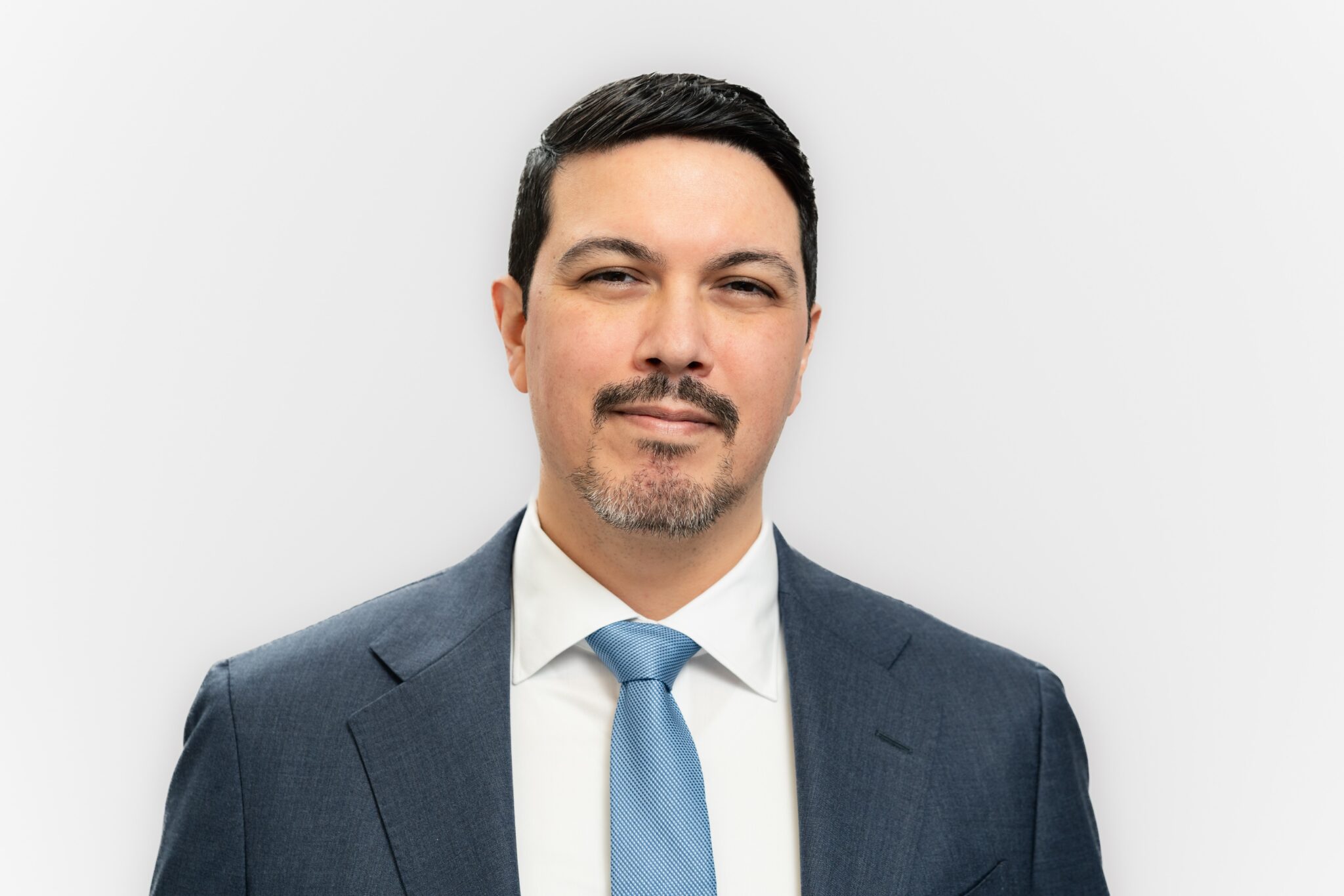 A professionally dressed man is shown in a studio-style headshot, centered against a plain light background and looking directly at the camera with a calm, slight smile. He wears a dark textured blazer, white dress shirt, and light blue tie, with neat dark hair and a closely trimmed goatee.