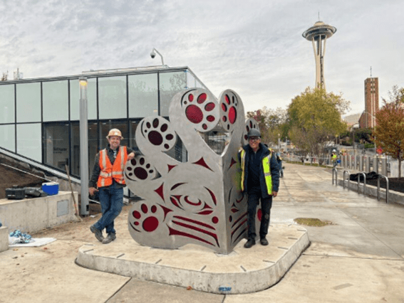 Photo of two people standing in front of a newly-installed piece of artwork. Both people wear reflective safety vests and hard hats. The Space Needle and other buildings are in the background.