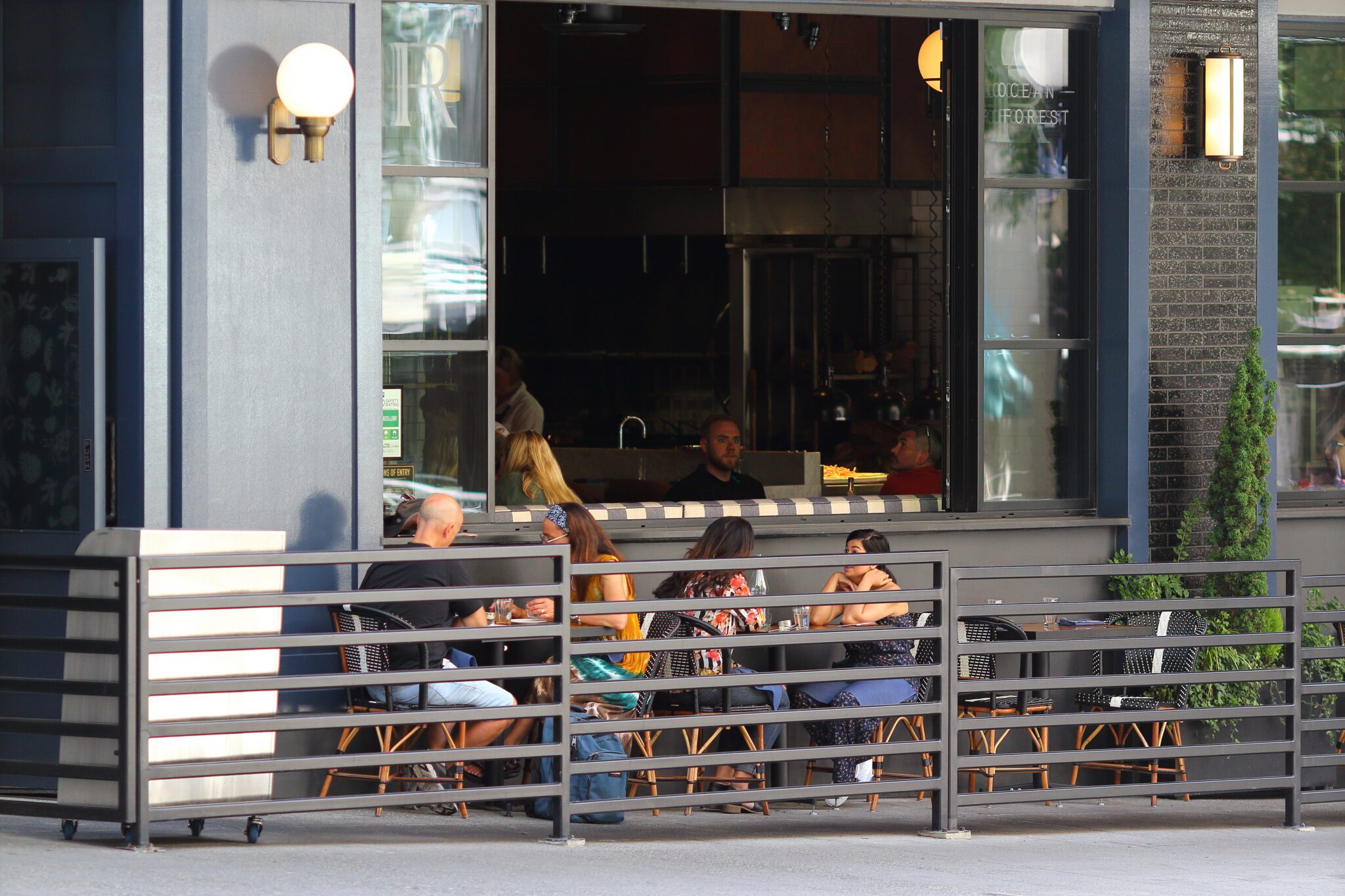 Four people, two at each table, sit at an outdoor table outside a dark, modern restaurant, separated from the sidewalk by a black metal fence. Through a large open window behind them, more people are seen sitting inside the dimly lit interior.