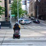 A person in a motorized wheelchair wearing a yellow jacket crosses a wide, striped crosswalk at a busy urban intersection. Nearby, a man looks at his phone while walking. The scene includes parked cars, traffic signals, road signs, and buildings on a sloping street.