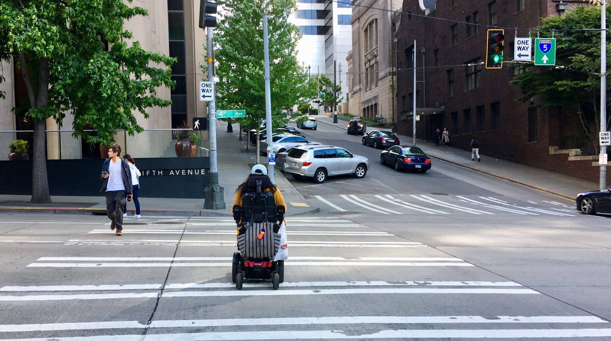 A person in a motorized wheelchair wearing a yellow jacket crosses a wide, striped crosswalk at a busy urban intersection. Nearby, a man looks at his phone while walking. The scene includes parked cars, traffic signals, road signs, and buildings on a sloping street.