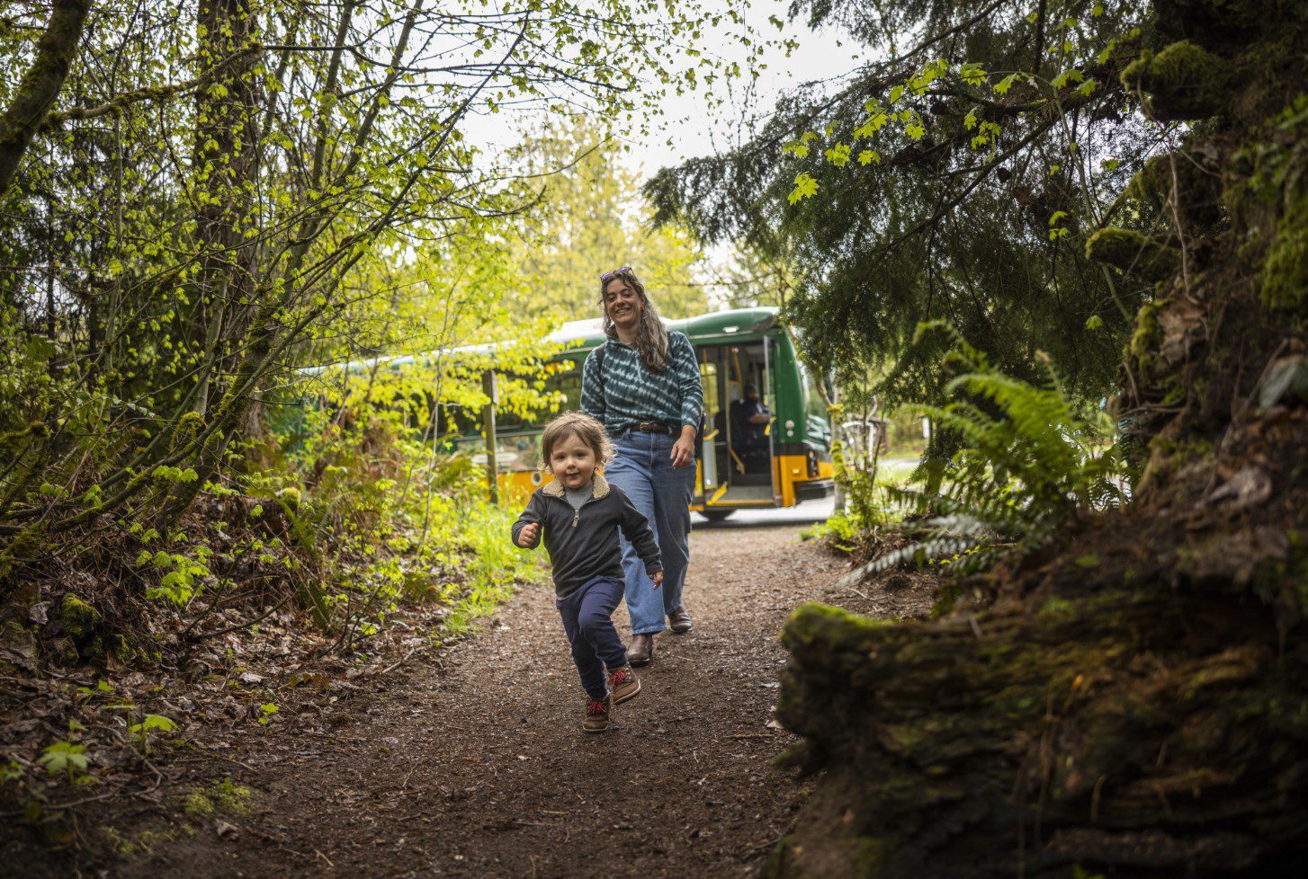 A smiling young child runs along a forest path while an adult follows close behind, with a green and yellow shuttle bus parked in the background. Tall trees, moss, and ferns surround the trail, creating a lush outdoor setting. The image conveys a joyful family outing and easy access to nature.