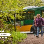 Three hikers walk down a forest trail after stepping off a green and yellow Trailhead Direct shuttle bus parked beside the path. Fresh spring leaves and ferns surround the scene, creating a lush backdrop that highlights the connection between transit and outdoor access. The bus branding and “Park. Ride. Hike.” message are visible, reinforcing that this is a promotional image for a hiking shuttle service.