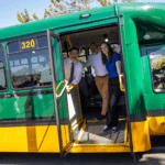 Three smiling people stand in the open doorway of a green-and-yellow shuttle bus marked Route 320 to Golden Garden, parked outdoors on a sunny day.