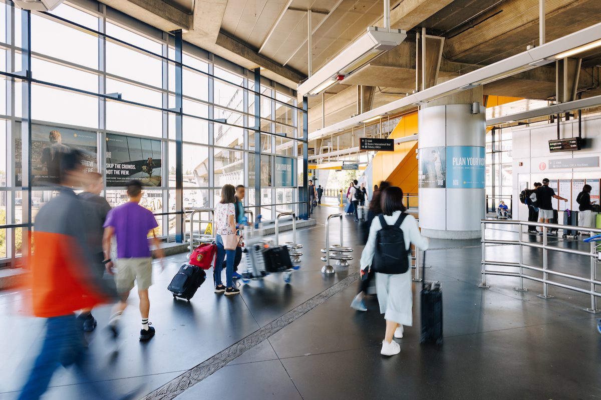 Travelers with luggage walk through a bright Link light rail station with ticket machines, and directional signs.